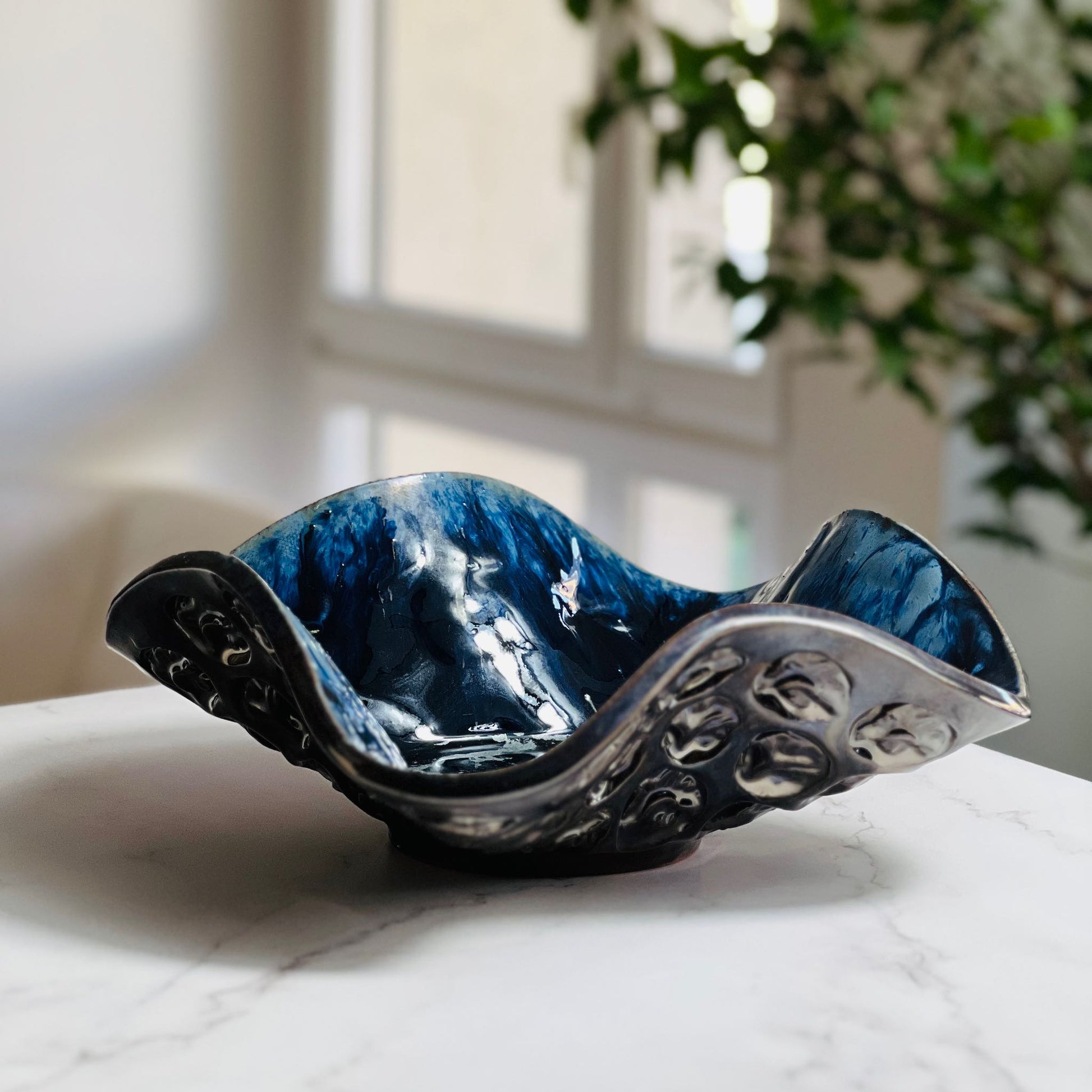 Handmade medium-sized decorative dark blue ceramic bowl with half-flower and tree texture displayed on a marble table with a living room background.